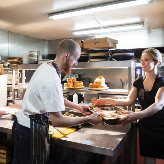 A caucasian male and female chef working together in a commercial kitchen. They are wearing kitchen clothing and aprons while the female is passing a tray of meat to her colleague.